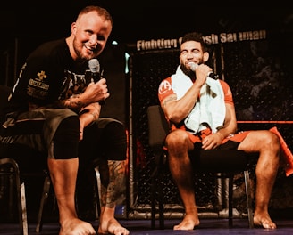 Two individuals are seated on a stage, each holding a microphone and smiling. They are in a sports-related setting with one wearing a casual black outfit and the other in sportswear, with a towel around his neck. The background includes elements of a fighting or martial arts cage.