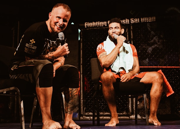 Two individuals are seated on a stage, each holding a microphone and smiling. They are in a sports-related setting with one wearing a casual black outfit and the other in sportswear, with a towel around his neck. The background includes elements of a fighting or martial arts cage.