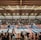A large indoor arena with a high ceiling, filled with an audience seated on bleachers. In the center, two fighters are engaged in a martial arts competition on blue and red mats. The scene is captured through a mesh fence, adding a textured foreground.