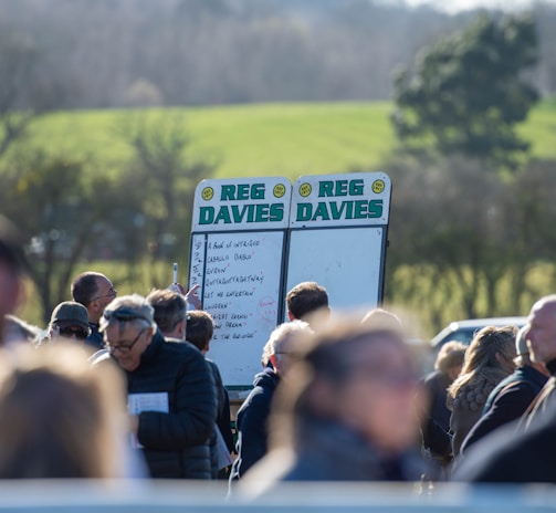 A group of people gathered outdoors with two large betting boards in the background labeled 'REG DAVIES'. The setting appears to be a countryside location with trees and fields visible in the distance. The crowd consists of individuals dressed in jackets and hats, engaging in conversation.