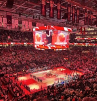 An indoor sports arena filled with spectators and a basketball court illuminated by red lighting. The scoreboards and video screens display promotional content, with numerous banners and championship flags hanging from the ceiling. Players and officials are gathered on the court, possibly in preparation for a game or event.