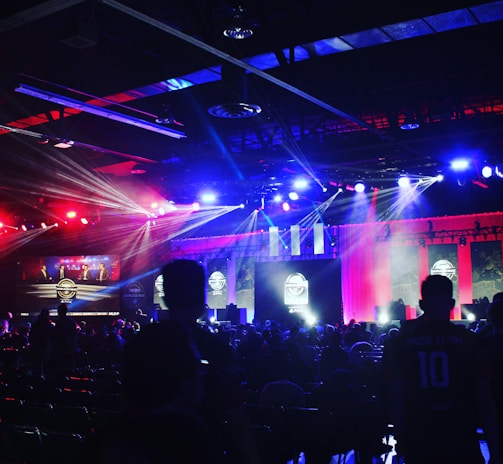 A dimly lit indoor arena filled with an audience. Bright stage lights in red and blue illuminate multiple large screens displaying graphics.