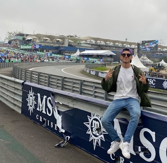 A person in casual attire sits on a barrier beside a racetrack, making peace signs with both hands. In the background, there is a stadium, a large crowd, promotional banners, and a screen displaying an advertisement. The scene has an overcast sky, and the setting appears to be a motorsport event.