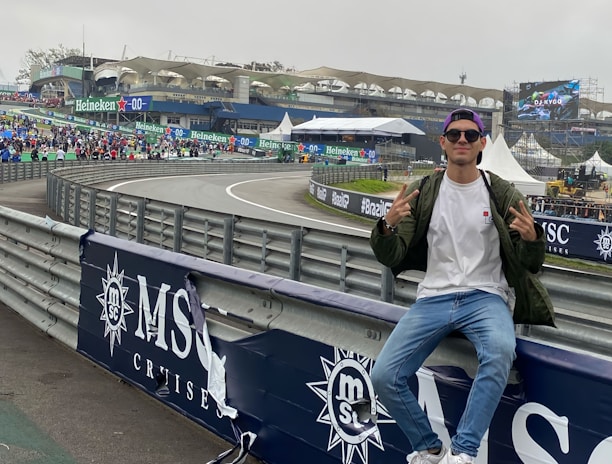 A person in casual attire sits on a barrier beside a racetrack, making peace signs with both hands. In the background, there is a stadium, a large crowd, promotional banners, and a screen displaying an advertisement. The scene has an overcast sky, and the setting appears to be a motorsport event.
