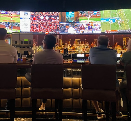 A group of people sit at a bar with their backs to the camera, watching multiple sports games displayed on large screens. The bar counter is lined with various bottles of liquor, and the atmosphere suggests a casual and relaxed social setting.