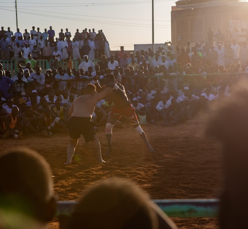 A vibrant wrestling match taking place outdoors, surrounded by a large crowd of spectators. The atmosphere is charged with excitement as people stand and sit around the dirt arena, intently watching the competitors. The sun is setting, casting a warm glow over the scene.