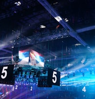 A large indoor arena is equipped with advanced lighting and audio systems. Suspended above the space are numerous digital displays showcasing numbers. The environment is filled with blue illumination and spotlights creating a vibrant atmosphere.
