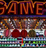 A brightly lit carnival game stall with a large 'GAMES' sign at the top surrounded by red and yellow lights. Various sports balls, including basketballs and soccer balls, are hanging from the ceiling and neatly arranged on shelves below.