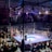 A crowded indoor arena with a boxing ring at the center, surrounded by an audience seated at tables. The scene is illuminated by overhead lights, casting a vibrant glow over the ring where two fighters are preparing for a match. Spectators are engaged, capturing the event with cameras and enjoying the lively atmosphere.
