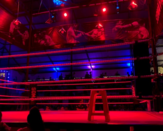 An empty boxing ring is illuminated by dramatic red and blue spotlights inside a dimly-lit arena. A small stool is placed in the center of the ring, surrounded by ropes. The arena is adorned with banners depicting action scenes, and a few silhouettes of spectators can be seen in the background.