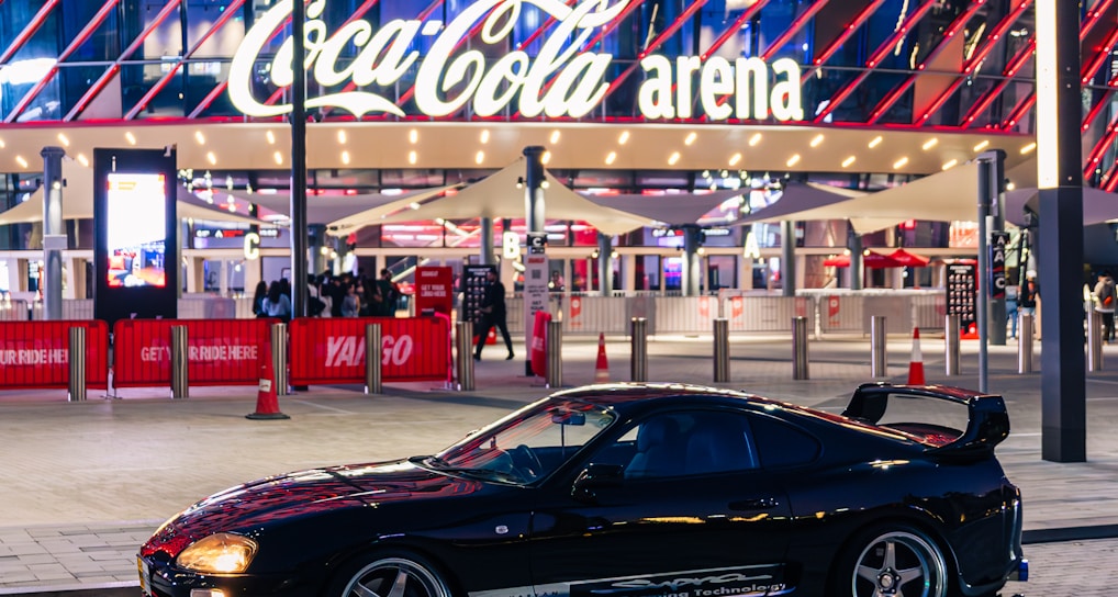 A sleek black sports car is parked in front of an illuminated arena entrance, with a large sign bearing the name of a famous beverage brand. The building features a modern, geometric facade with bright red and blue lights, creating a vibrant and lively atmosphere.